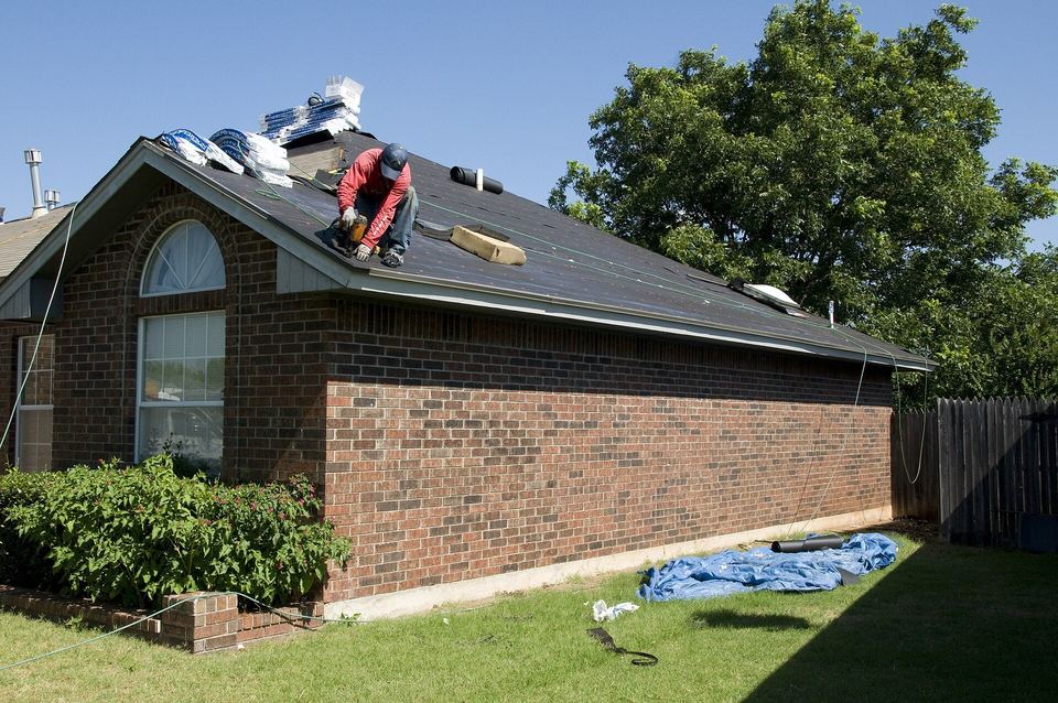 Roofer kneeling on a brick ranch house roof, nailing new asphalt shingles into place