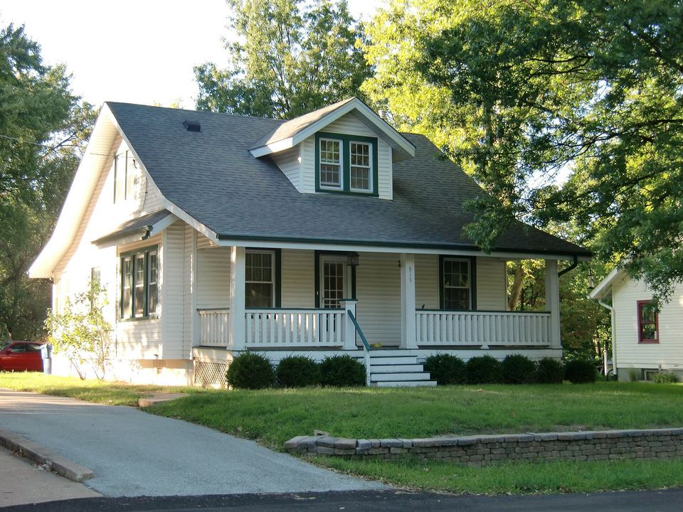 White bungalow with a dark gray shingle roof, covered front porch and green lawn