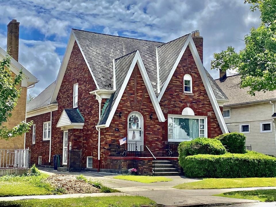 Red brick Tudor-style house with steep gray shingle gables and trimmed front hedges