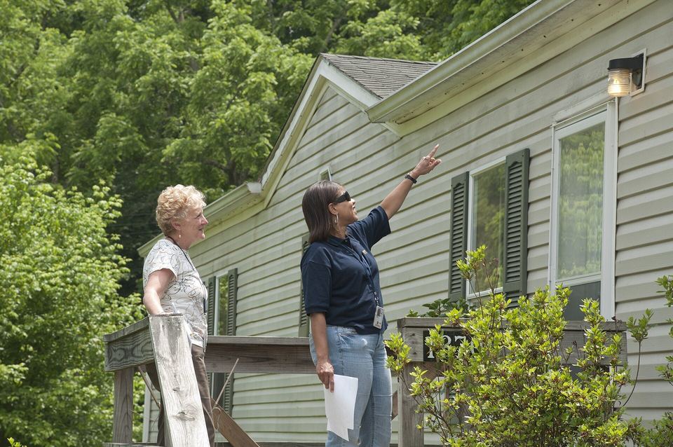 Inspector holding paperwork points at a house roofline while a homeowner looks on