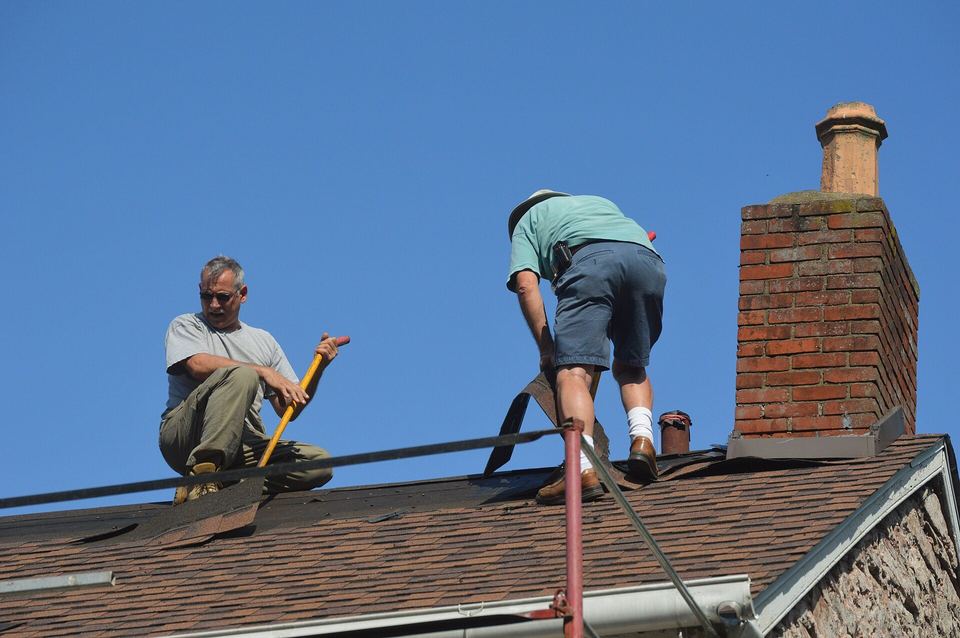 Two roofers working on a shingle roof beside a brick chimney under blue sky
