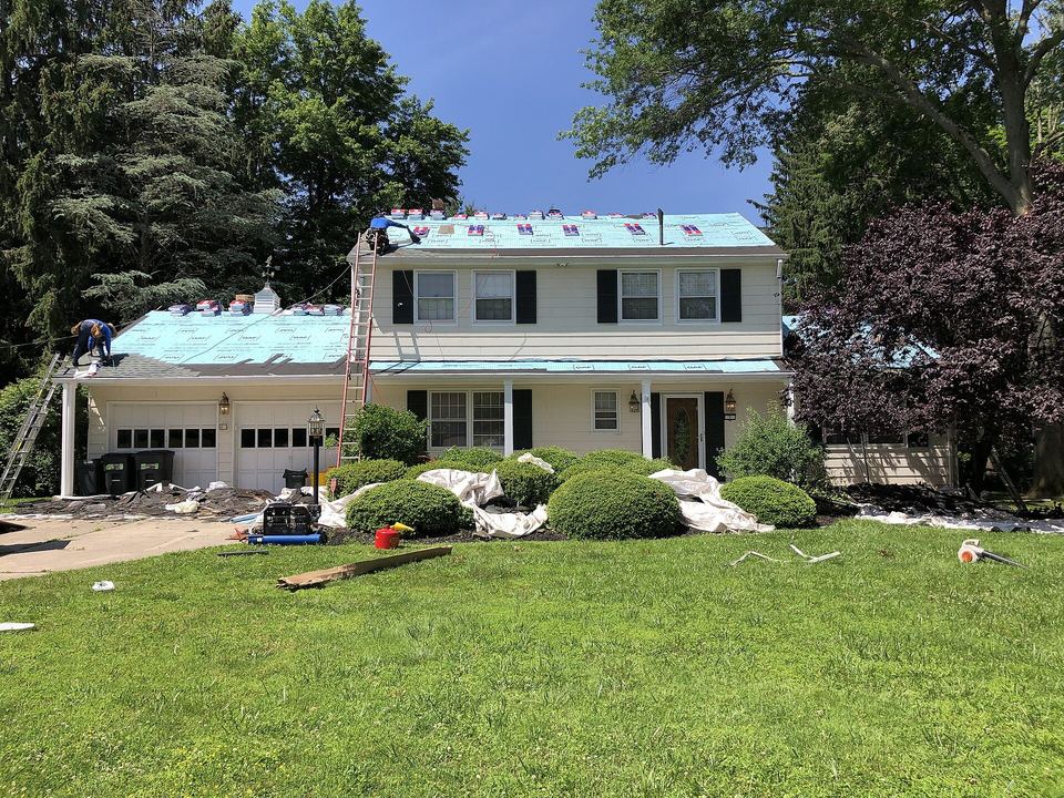 Two-story house with light blue synthetic underlayment covering the roof during replacement